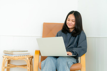 Portrait, a cute Asian teenage girl. wearing gray shirt sitting on a chair with a notebook computer in the bedroom to study online with a happy face