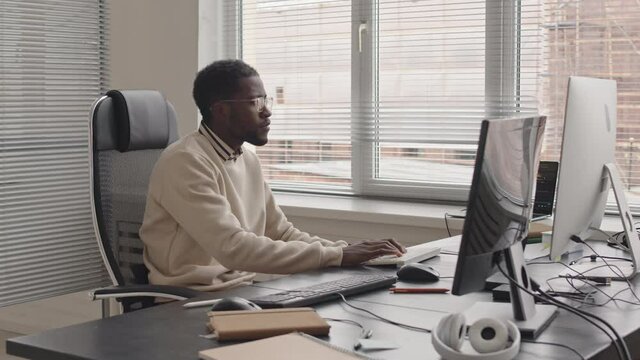 Stab shot of young African-American software developer working on pc sitting at desk by window in bright modern office