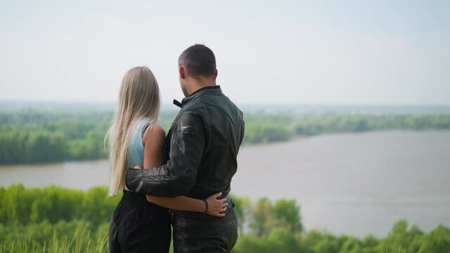 Long haired blonde woman and motorcyclist in protective suit hug looking at tranquil river from high hill slope on spring day backside view slow motion