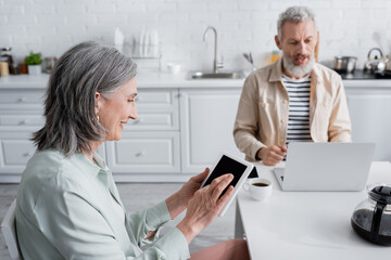 Smiling mature woman using digital tablet near blurred husband with laptop and coffee in kitchen.