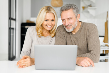 Happy middle-aged couple spends leisure time with a laptop at home. Senior spouses sit at the desk in kitchen, watching video, web surfing or shopping online together, look at screen and smile