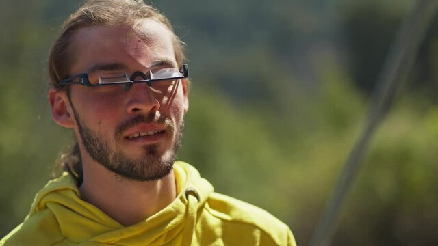 Young Attractive Man Rock Climber In The Mountains Belaying Climbing Partner And Setting Up Rope, Healthy Lifestyle And Fitness. Man Looking Up Through Specialized Climbing Eyeglesses With Prisma