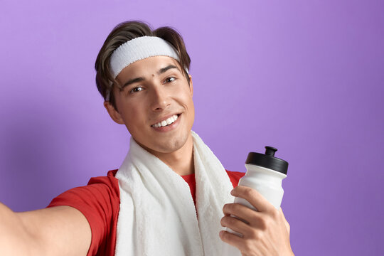 Close up shot of a smiling young sportsman taking selfie with smartphone, holding gym water bottle in hand, wearing red t-shirt, towel over neck and white headband, isolated on violet background.
