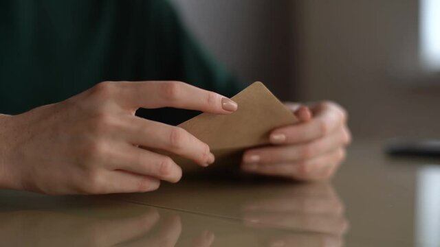 Close-up hands of unrecognizable young woman putting handwritten Christmas letter into envelope and closes it, sitting at table at home, selective focus.