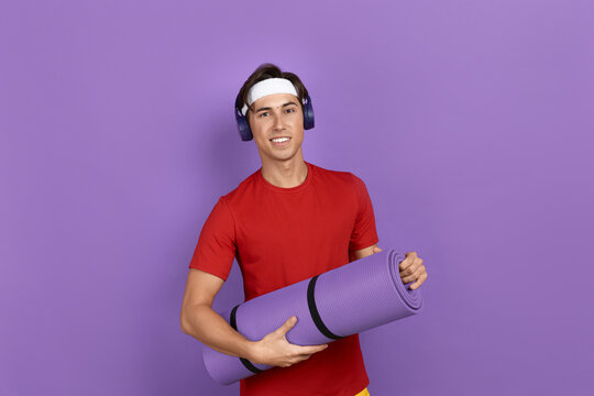 Young smiling athlete or coach is holding fitness or yoga mat with hands, leaving sport club after training, wearing red t-shirt, blue headphones and white headband, isolated on violet background.