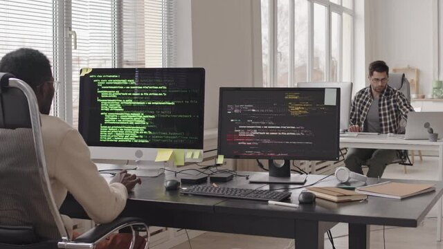 Stab shot of team of programmers sitting at workplaces in front of computer monitors with running program codes on screens in modern office