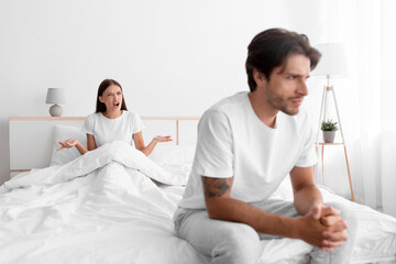 Unhappy frustration european young man ignoring angry woman, lady sits in bed and scream on guy in white bedroom