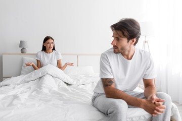 Fototapeta premium Sad depressed young european husband with stubble looks at screaming wife in bedroom interior, empty space