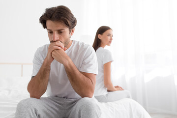 Sad upset offended millennial caucasian man sitting on bed, ignoring woman in bedroom interior