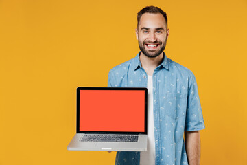 Young smiling happy freelancer copywriter satisfied man 20s wear blue shirt hold use work on laptop pc computer with blank screen workspace area isolated on plain yellow background studio portrait