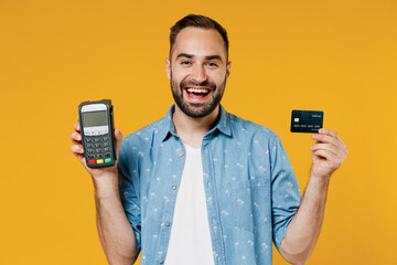 Young smiling fun happy man 20s wearing blue shirt white t-shirt hold wireless modern bank payment terminal to process acquire credit card payments isolated on plain yellow background studio portrait