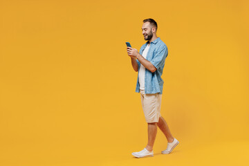 Full body young smiling happy caucasian man 20s wearing blue shirt white t-shirt hold in hand use mobile cell phone walk isolated on plain yellow background studio portrait. People lifestyle concept.