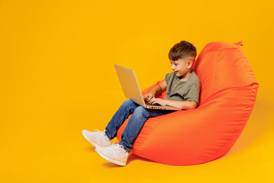 Full Body Little Small Happy Boy 6-7 Years Old Wear Green T-shirt Sit In Bag Chair Hold Use Work On Laptop Pc Computer Isolated On Plain Yellow Background Mother's Day Love Family Lifestyle Concept