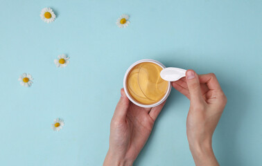 Hand holds a jar of gold collagen patches on a blue background with chamomile