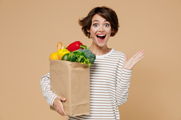 Young excited happy fun vegetarian woman 20s in casual clothes hold paper bag with vegetables bell pepper after shopping in greengrocery spread hands isolated on plain pastel beige background studio
