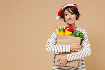Young fun woman 20s in casual clothes christmas hat hold paper bag with vegetables look aside on...