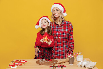 Two woman mother fun child baby girl in red Christmas hat isolated on plain yellow wall background studio. Mom little kid cook ginger cookie at kitchen table home. Happy New Year 2022 holiday concept