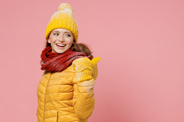 Promoter smiling fun young woman 20s years old wears yellow jacket hat mittens pointing back behind on workspace area copy space mock up isolated on plain pastel light pink background studio portrait.
