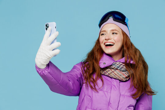 Skier smiling woman in warm purple padded windbreaker jacket ski goggles mask spend extreme weekend in mountains do selfie shot mobile cell phone post photo isolated on plain blue background studio