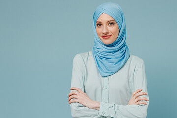 Young smiling confident arabian asian muslim woman in abaya hijab hold hands crossed folded isolated on plain blue color background studio portrait. People uae middle eastern islam religious concept.