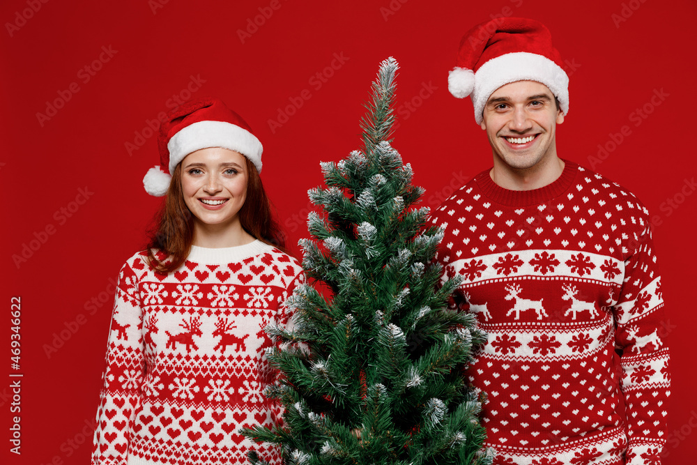 Wall mural Young fun couple friends two man woman 20s in sweater hat hold fir-tree looking camera isolated on plain red background studio portrait. Happy New Year 2022 celebration merry ho x-mas holiday concept.