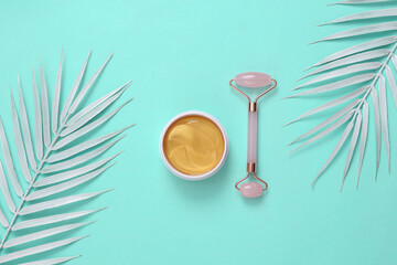 Facial massage roller with gold cream or patches in a jar, palm white leaves on a turquoise background. Beauty, spa salon concept