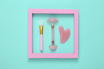 Face roller, brush and gua sha stone massager in pink frame on blue background. Top view