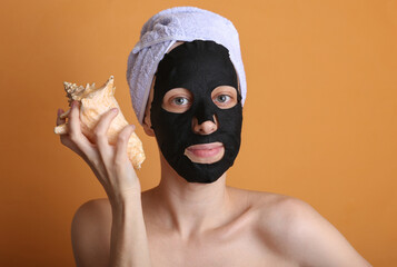 Beautiful woman in a cleansing black mask with a towel on her head holding seashell on orange background