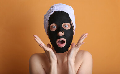 Shocked beautiful woman in a cleansing black mask with a towel on her head on an orange background
