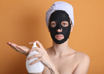 Lovely woman in a cleansing black mask with a towel on her head applies cream to her hands on an orange background