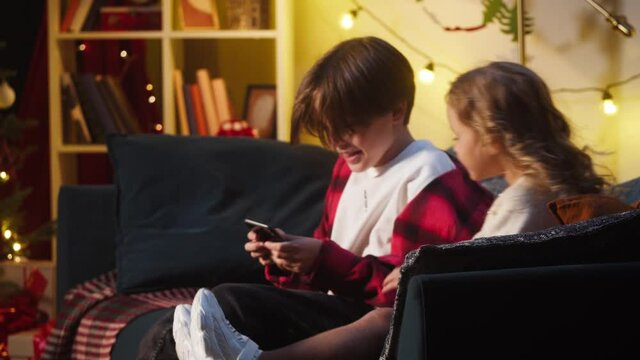 Children running to sofa, using smartphone together in living room. Brother and sister having fun at home. Free time during christmas holidays. New year atmosphere, happy childhood.
