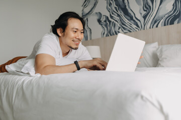 Happy asian man work with laptop on the bed in the hotel. Concept of workation.