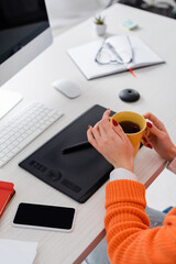 high angle view of cropped woman holding cup of tea near graphic tablet, keyboard and blurred monitor