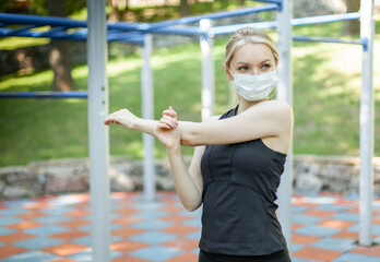 Young fit woman doing hand stretching in medical mask on workout site