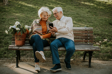 Senior couple sitting on the benchwith basket full of flowers and lookin at mobile phone