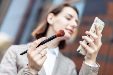 Young stylish businesswoman applies makeup with a brush and looks in the mirror against the...