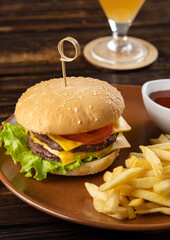 Homemade burger with juicy beef patty, tomatoes, cheese, sauce, lettuce leaves, French fries on a round plate on a dark wooden background. Fast food. Vertical orientation, close-up, no people