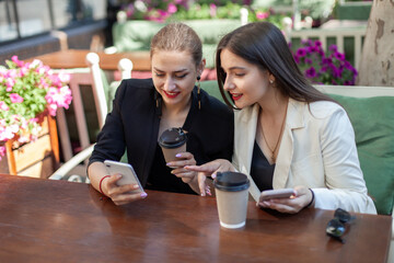 Two cute women friend are looking at screen smartphone while sitting at table in an outdoor cafe