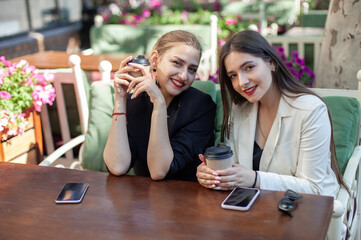 Two smiling young caucasian women with coffee cups are sitting at table in outdoor cafe