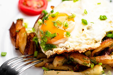Fried potatoes with egg with liquid flowing yolk, herbs and tomatoes on a white plate.