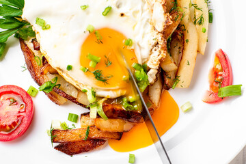 Fried potatoes with egg with liquid flowing yolk, herbs and tomatoes on a white plate.