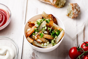 Fried potatoes in a white plate with green onions, ketchup on a white wooden background.