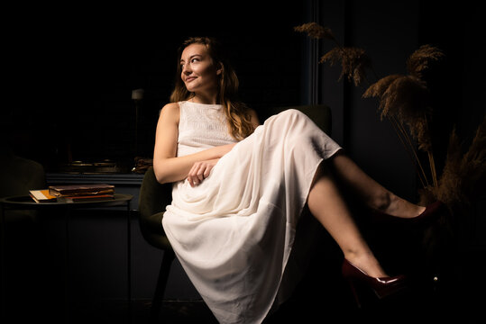 A young woman in a white dress sits relaxed on an armchair in a dark room.