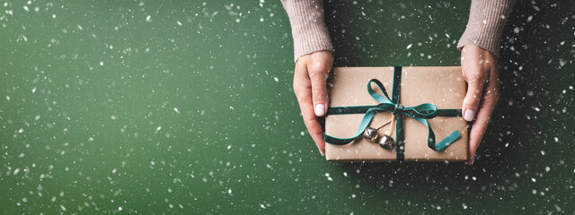 Snowy banner with female hands holding Christmas gift box tied velvet ribbon on a green background.