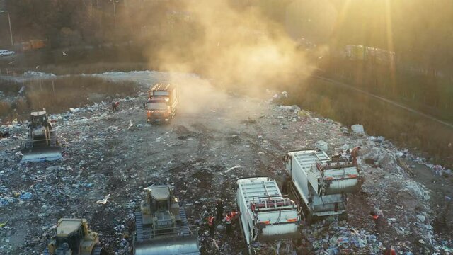 Aerial view of a garbage trucks unload garbage at a city dump at sunset. Bulldozers crush and level debris. Flocks of birds are circling over the dump.
