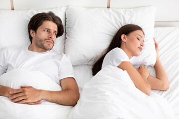 Disgruntled sad caucasian young guy does not sleep, lies in bed, looks at lady, woman snores in white bedroom
