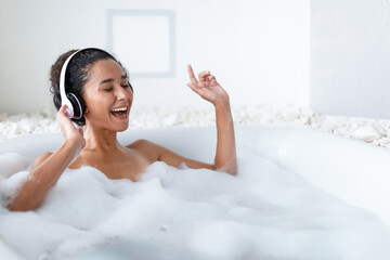 Attractive young lady wearing headphones and singing while lying in hot bubbly bath at home, copy...