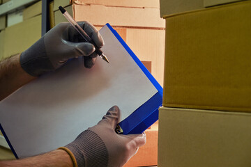 Men gloved hands write with a pen on a tablet with paper, checking a warehouse with goods in boxes
