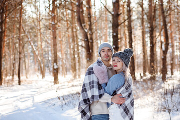 Fototapeta na wymiar Happy couple wearing warm clothes and checkered plaid hugging in winter snowy forest, copy space.