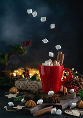 Hot chocolate with marshmallows in a red mug on dark wooden boards. Marshmallows are hanging in the air and scattered on the table. Fir branches, cinnamon tubes, cookies and lights in the background